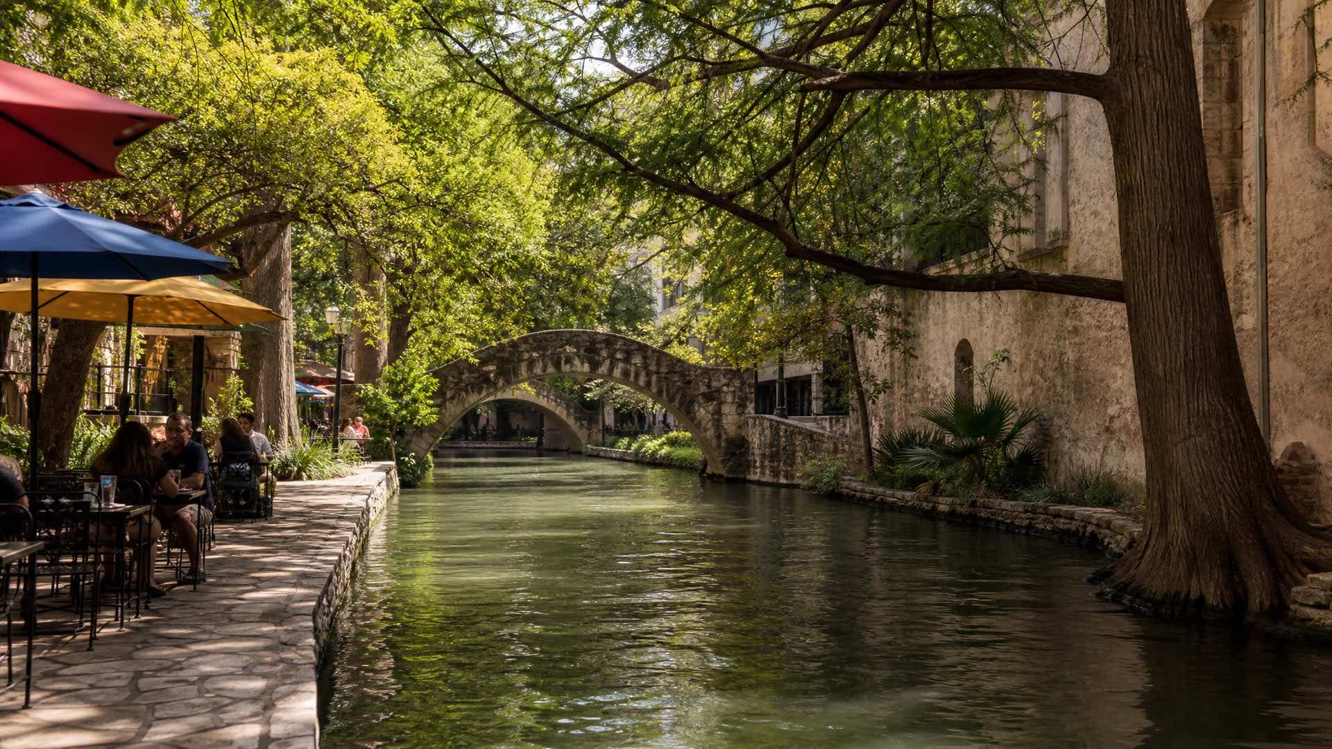 A stone walkway and arched footbridge along the green San Antonio River below street level, cafe umbrellas under cypress shade