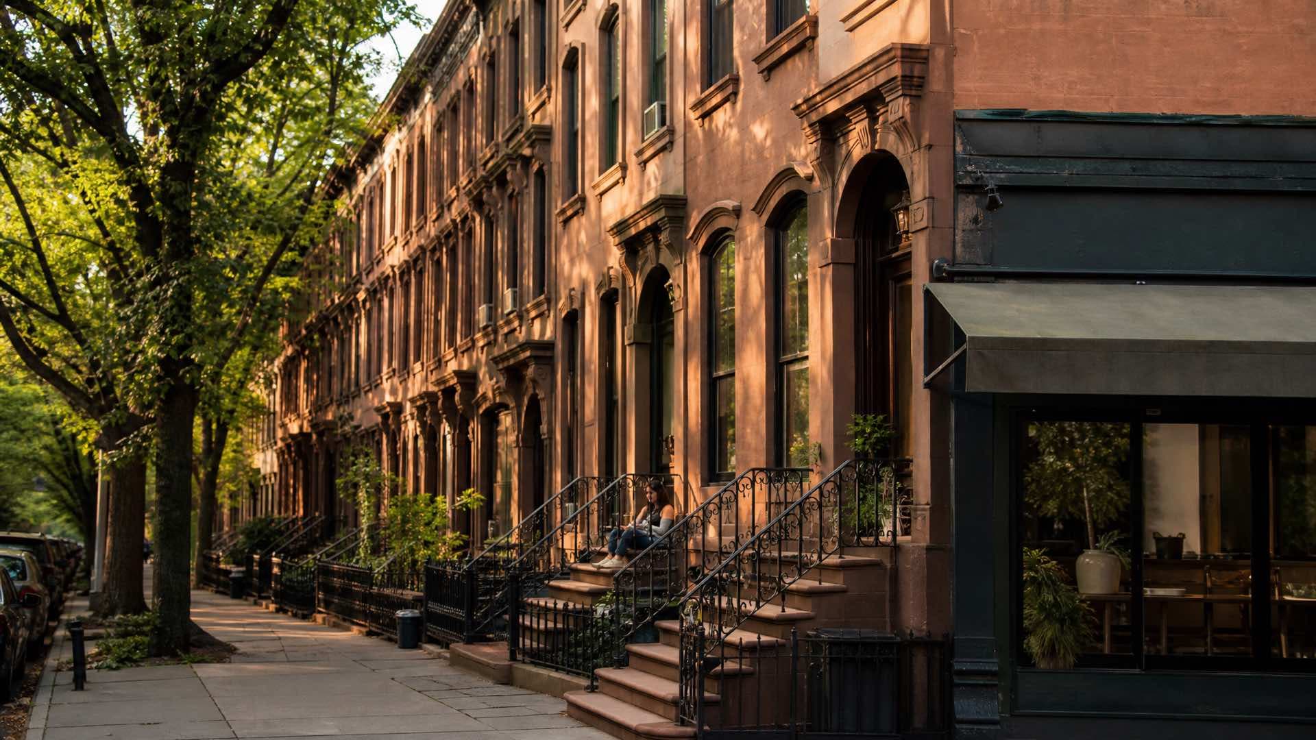 A Brooklyn brownstone block of stoops and iron railings under mature street trees, a corner shopfront at the end of the row