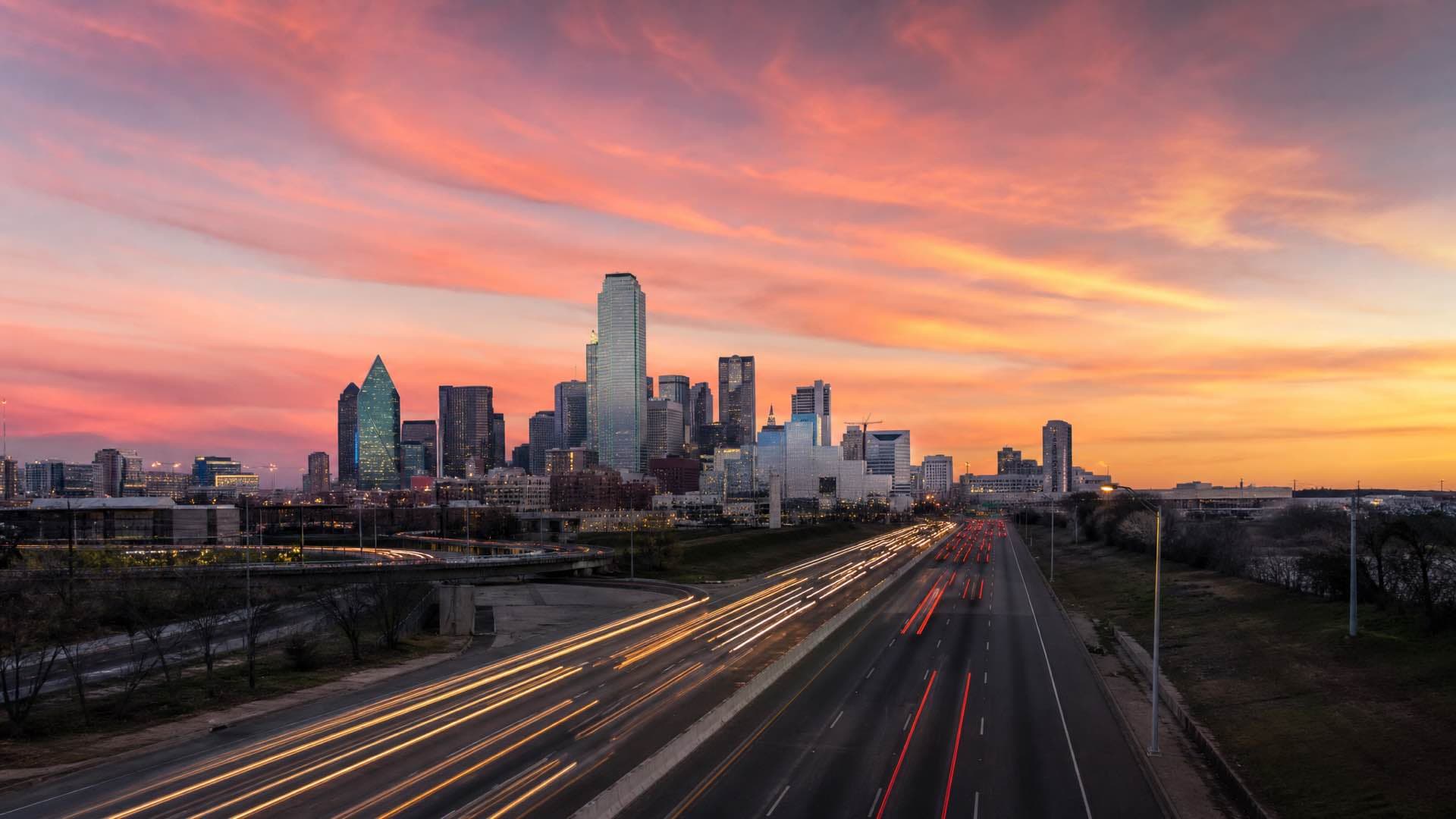 The Dallas skyline at dusk seen across a wide freeway, glass towers under an enormous Texas sky