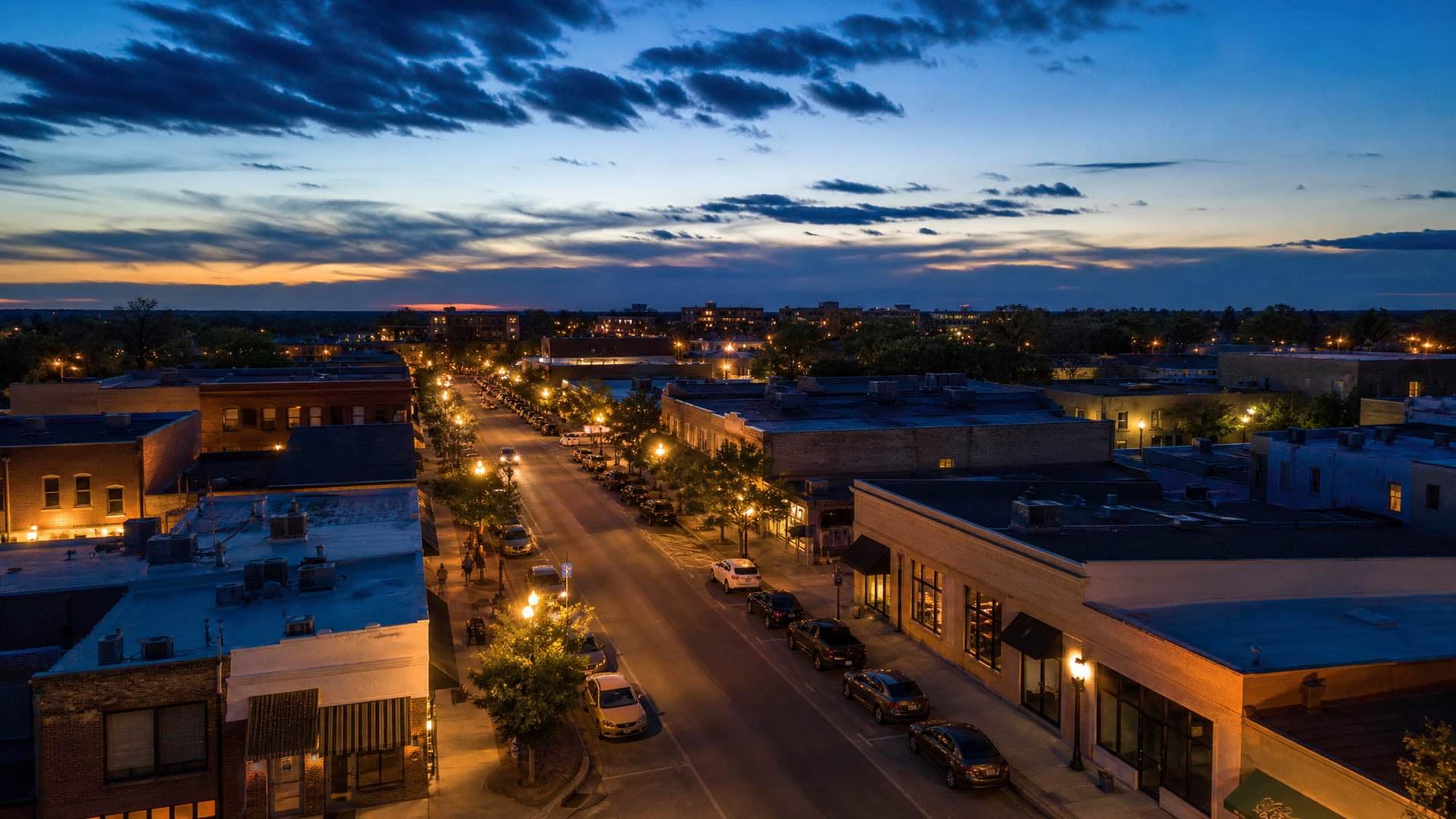 An aerial view at dusk of an American mixed commercial district, low-rise storefronts along a street grid with streetlights coming on