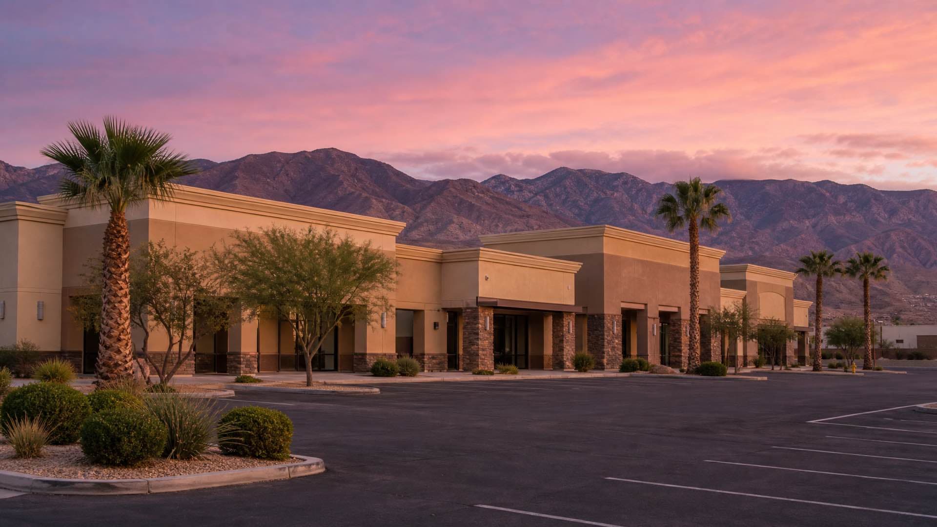 A low desert-modern commercial plaza in the Las Vegas valley at dusk, dry mountains ringing the horizon