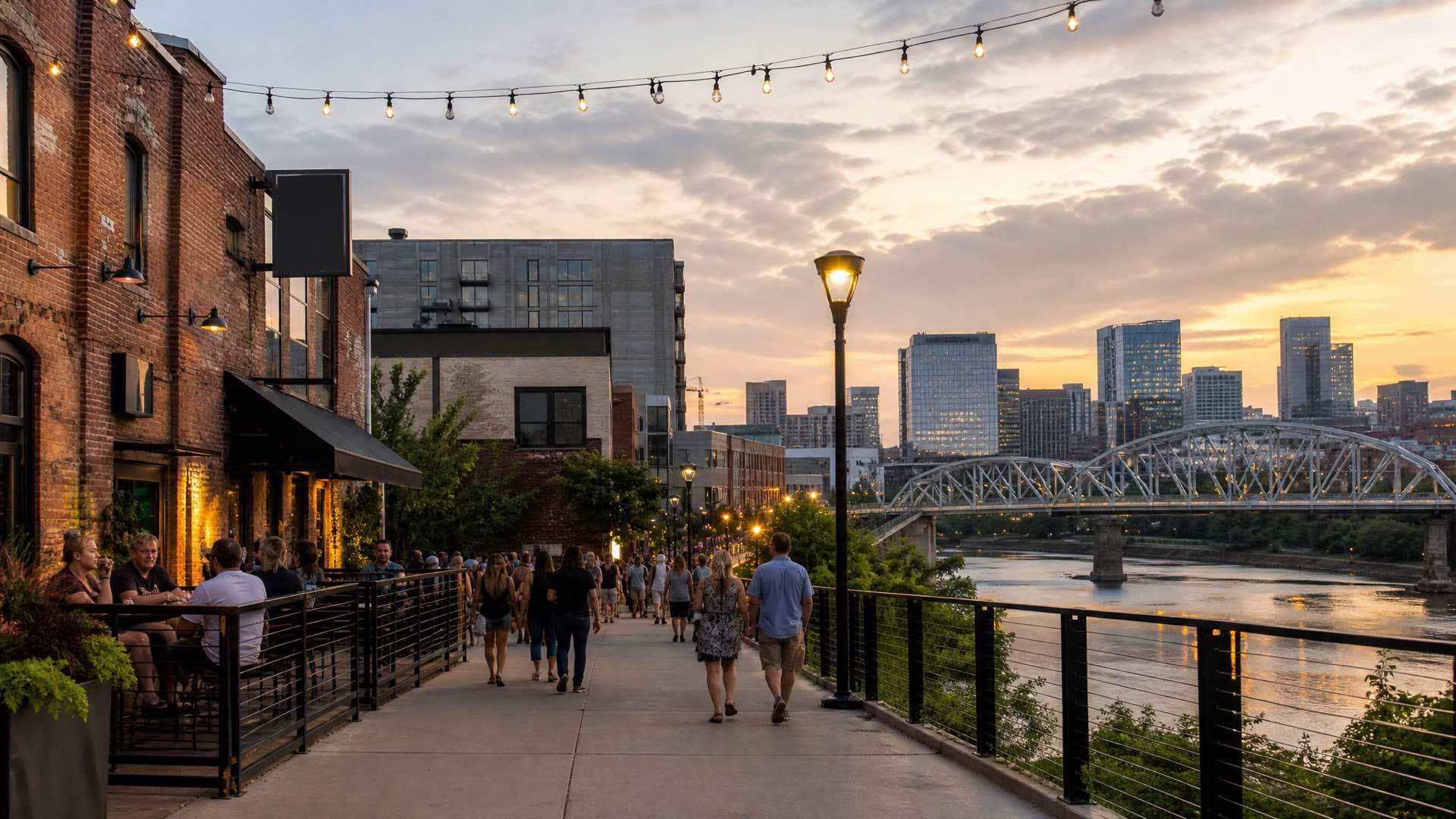 Converted red brick warehouses in a Nashville district at warm dusk, a steel pedestrian bridge over the river beyond