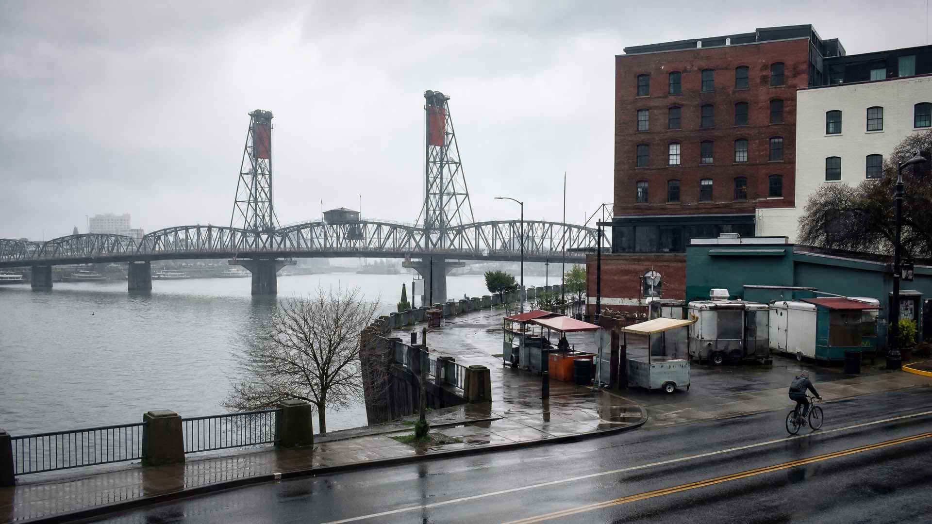 A cyclist on a wet Portland waterfront road, food carts and brick warehouses beside a steel lift bridge under grey sky